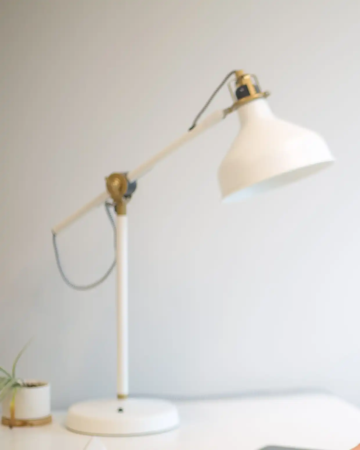 A white desk lamp with a conical shade, brass joints and its cable running outside the arm, on a white desk